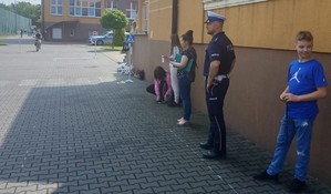 Policjant stoi przy murze budynku szkoły obok kilkoro dzieciaków, w oddali chłopiec na rowerze.