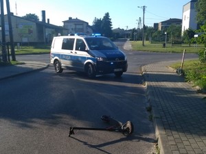 Na zdjęciu widać skrzyżowanie ulic, w poprzek ulicy zaparkowany policyjny radiowóz oznakowany, przed nim przewrócona na ulicy hulajnoga.