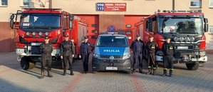 Przed budynkiem Komendy Państwowej Powiatowej Straży Pożarnej w Pajęcznie stoją dwa wozy strażackie, radiowóz, policjant, policjantka, 4 strażaków.