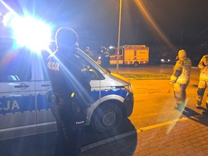 Noc, radiowóz oznakowany policyjny, obok policjant w mundurze, w oddali dwóch umundurowanych strażaków.