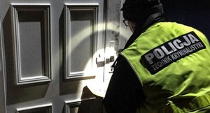 Policjant w kamizelce z napisem policjant techniki kryminalistyki, podświetla latarką drzwi.